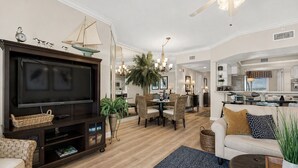 A modern living room with a flat-screen TV, a dining area with chairs, a kitchen in the background, and a ceiling fan. - Beachfront 2br/2ba condo on the 3rd floor with free beach chairs included! (Panama City Beach)