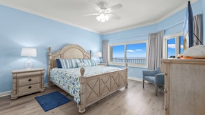 Quarto com cama, mesa de cabeceira, abajur, cadeira e vista para o oceano pela janela.