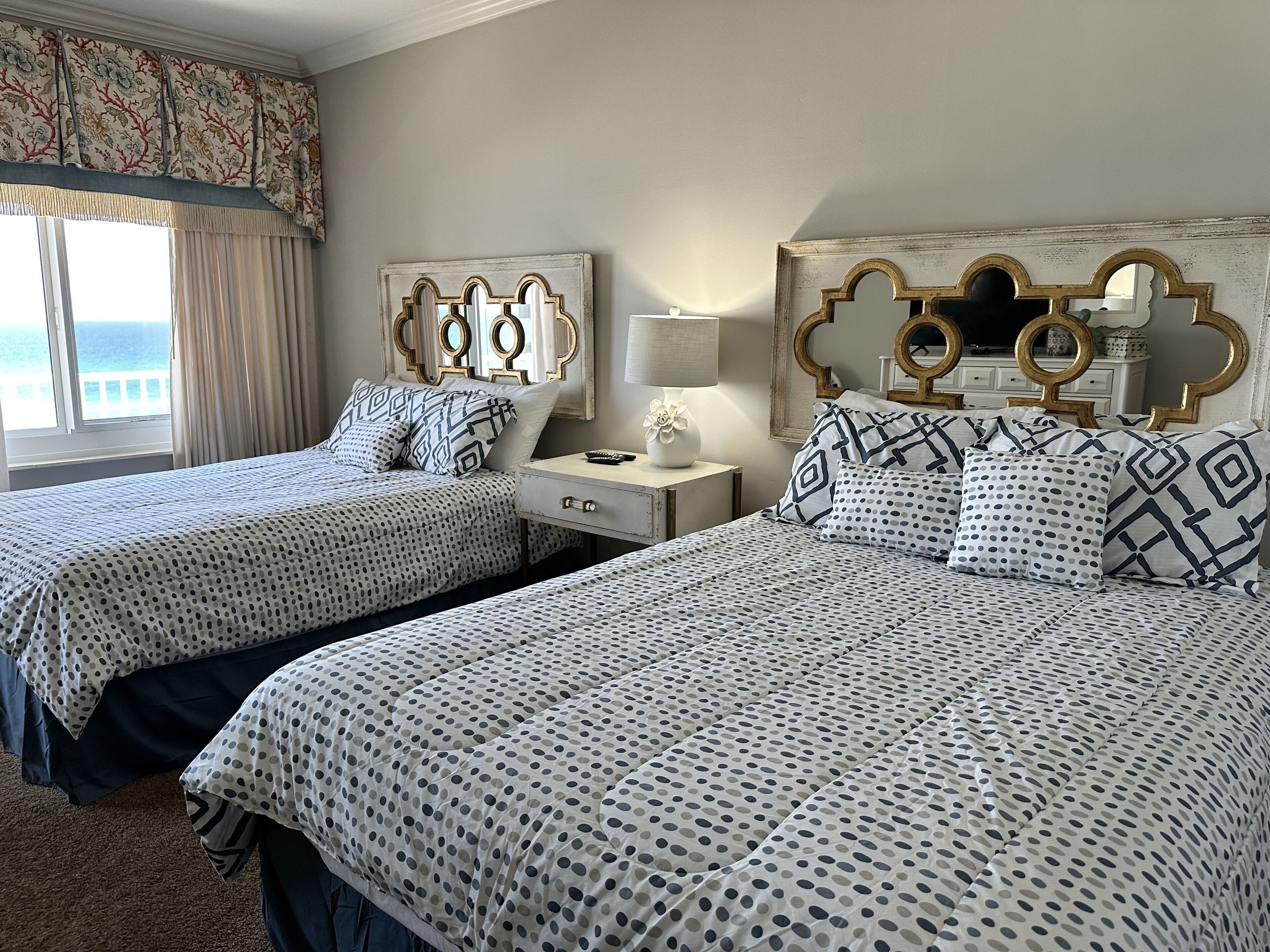 Een hotelkamer met twee bedden, elk met een bekleedsel met stippen. Er staat een nachtkastje met een lamp en een bloemenvaas. De kamer heeft een groot raam met gordijnen en uitzicht op de oceaan.