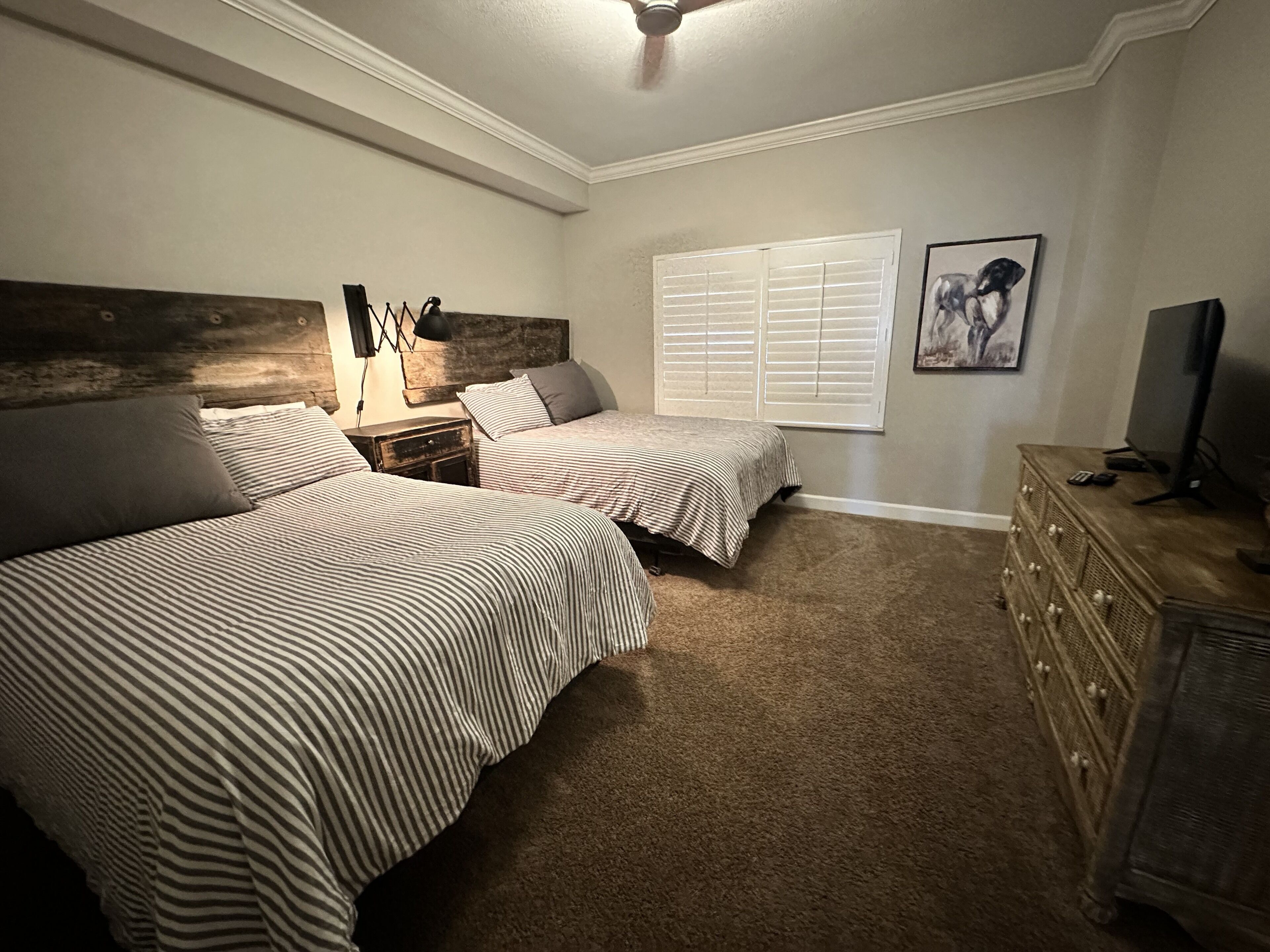 A bedroom with two beds, a wooden headboard, a nightstand, a dresser, a television, and a window with blinds.