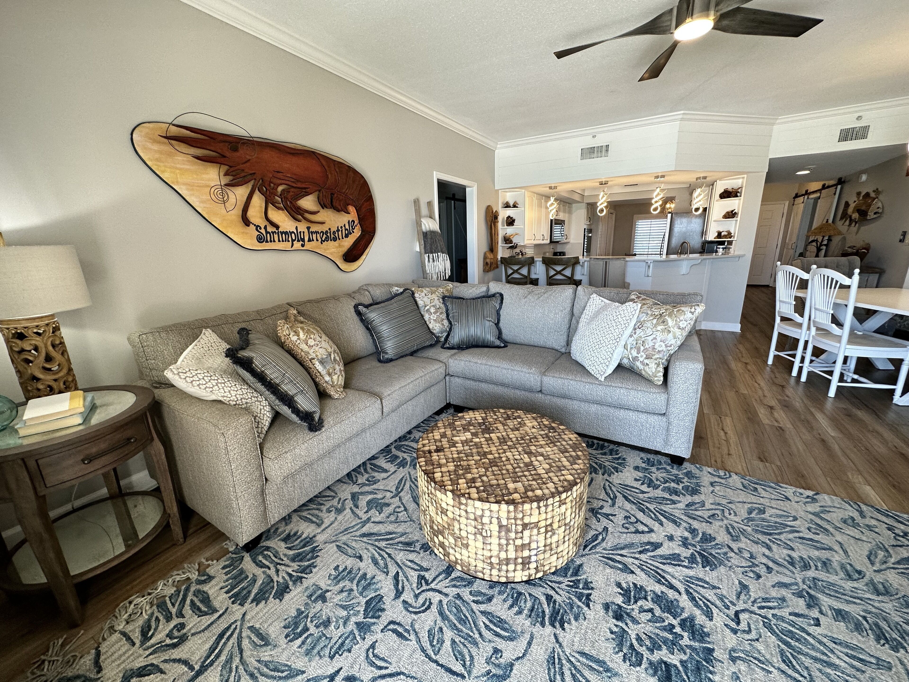 A living room with a sectional sofa, a round woven ottoman, a wall-mounted decorative piece, and a dining area with a table and chairs.
