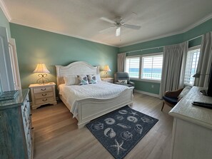 Quarto com cama, duas mesas de cabeceira, ventoinha no teto, janela com cortinas e vista para o mar.