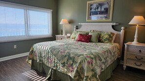 A bedroom with a floral bedspread, a view of the ocean through the window, and a painting on the wall.