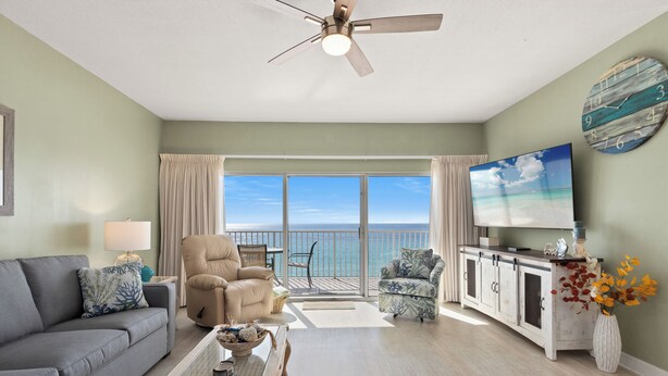 A spacious living room with a grey sofa, a beige armchair, a coffee table with decorative items, a television, and a view of the ocean through large windows.