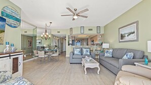 A spacious living room with a grey sofa, a white coffee table, and a dining area with wicker chairs. A ceiling fan and chandelier are visible.