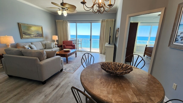 A spacious living room with a large sofa, a dining table with chairs, a ceiling fan, and a view of the ocean through large windows. - Coral Reef Unit #1405 Beachfront 2 bedroom 2 bath condo on the TOP floor!!! (Panama City Beach)
