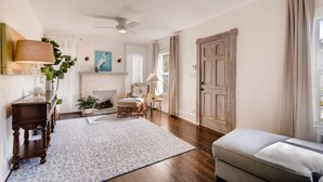 A spacious living room with a fireplace, wooden flooring, and a large window with curtains. - The Periwinkle House 4bd 2ba Sleep 8 (West Palm Beach)