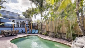 A swimming pool with a curved edge, surrounded by a wooden fence and palm trees. - The Periwinkle House 4bd 2ba Sleep 8 (West Palm Beach)
