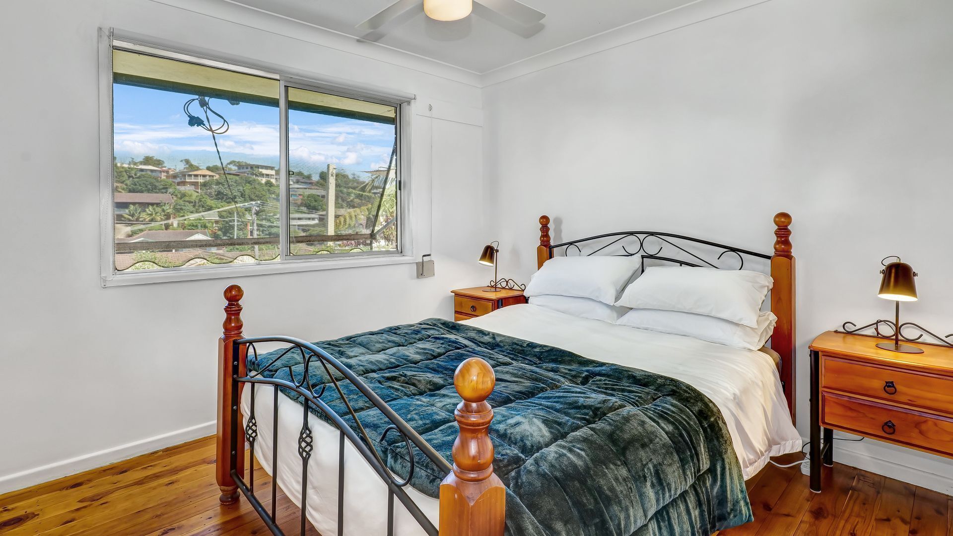 Une chambre à coucher avec un lit, une tête de lit en bois, une table de chevet, une lampe et une fenêtre donnant sur des bâtiments et de la verdure.