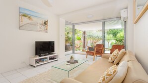 A modern living room with a glass coffee table, a beige sofa, and a flat-screen TV on a white stand. - Sawtell Beachside on 4th (Sawtell)