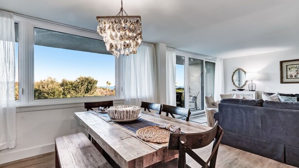 A dining area with a wooden table, chairs, and a chandelier. There is a large window with a view of trees and a clear sky.