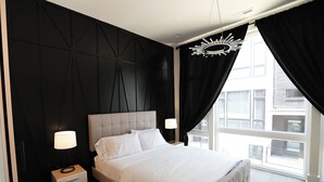 A modern bedroom with a large bed, a nightstand, a wall-mounted lamp, and a view of the outside through a window with black curtains. - Luxe Nashville 3-bedroom Townhouse w/rooftop deck (Nashville)