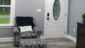 Una habitación con un sillón estampado, un sofá de cuero, un ventilador de techo, una puerta con un panel de vidrio decorativo y una ventana con vista a árboles.
