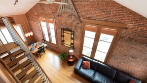 Un espace de vie de style industriel, avec des murs en briques apparentes, un escalier en bois, un canapé en cuir noir et un coin repas comprenant une table et des chaises.
