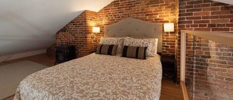 A bedroom with a sloped ceiling, exposed brick walls, a large bed with patterned bedding, and two bedside lamps.