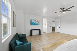 A spacious living room with a teal sofa, a wooden floor, a ceiling fan, and a painting on the wall.