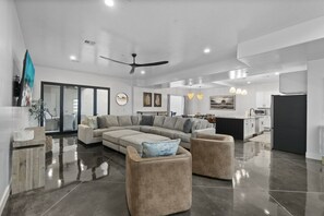 A modern living room with a sectional sofa, two armchairs, a ceiling fan, and a kitchen area in the background.
