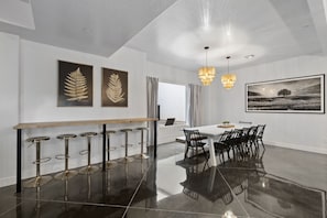 Ein moderner Essbereich mit einem langen Tisch, Barhocker, einer Chandelierleuchte und gerahmten Kunstwerken an den Wänden.