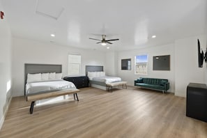Ein geräumiges Zimmer mit zwei Betten, einer Bank, einem grünen Sofa, einem Deckenventilator und einem großen Fenster.