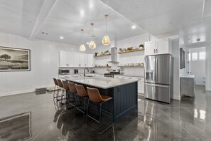 Una cocina moderna con una isla central, taburetes de bar, electrodomésticos de acero inoxidable, iluminación colgante y un amplio ventanal.