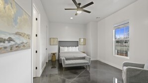 A bedroom with a bed, a grey sofa, a ceiling fan, a window with a view, and a painting on the wall. - Brand New Home! Roof Top Deck, Games for everyone, Private Pool (Hurricane)