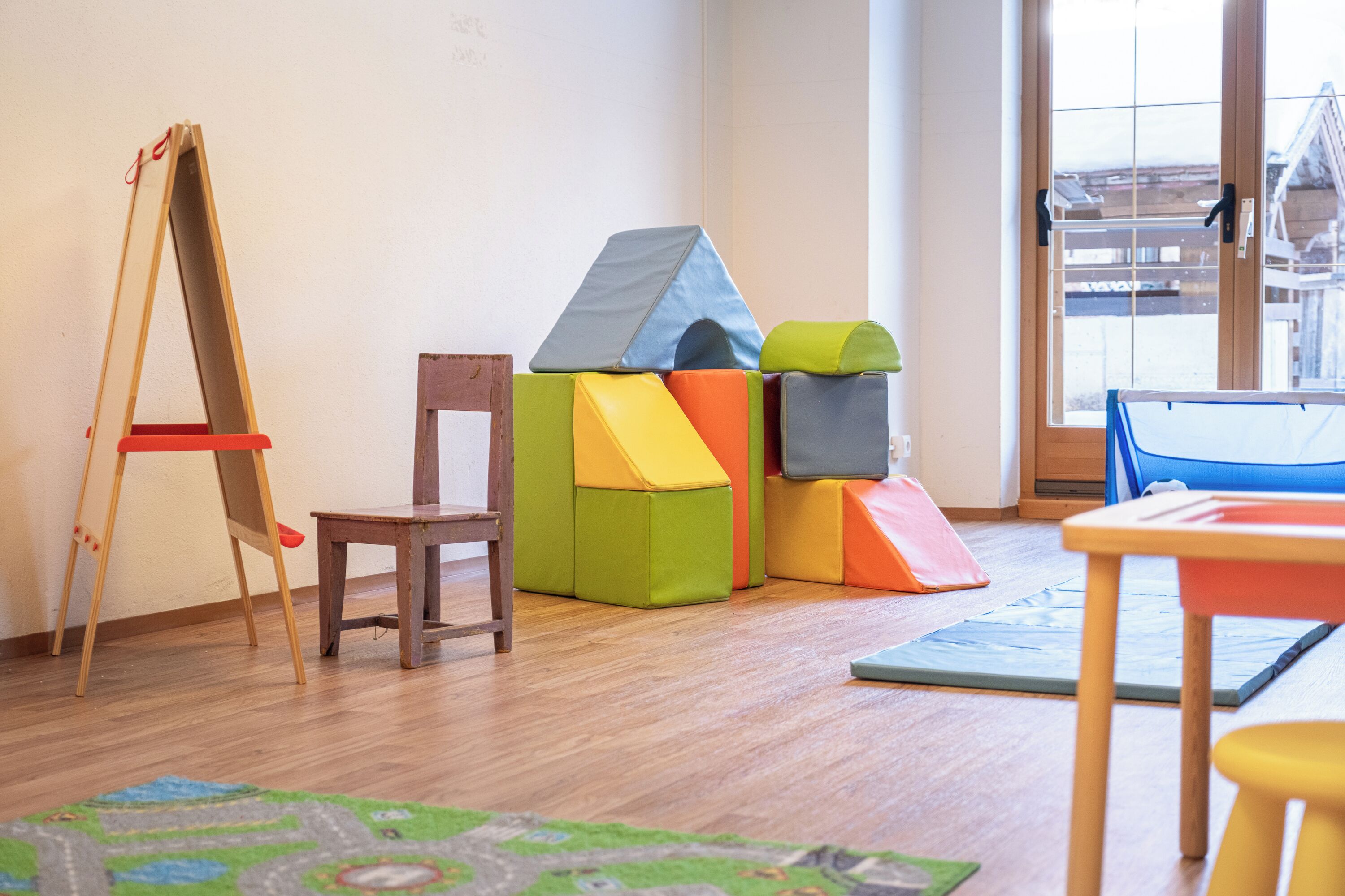A room with a wooden floor, a wooden chair, a small table, colorful cushions, and a view through a window.