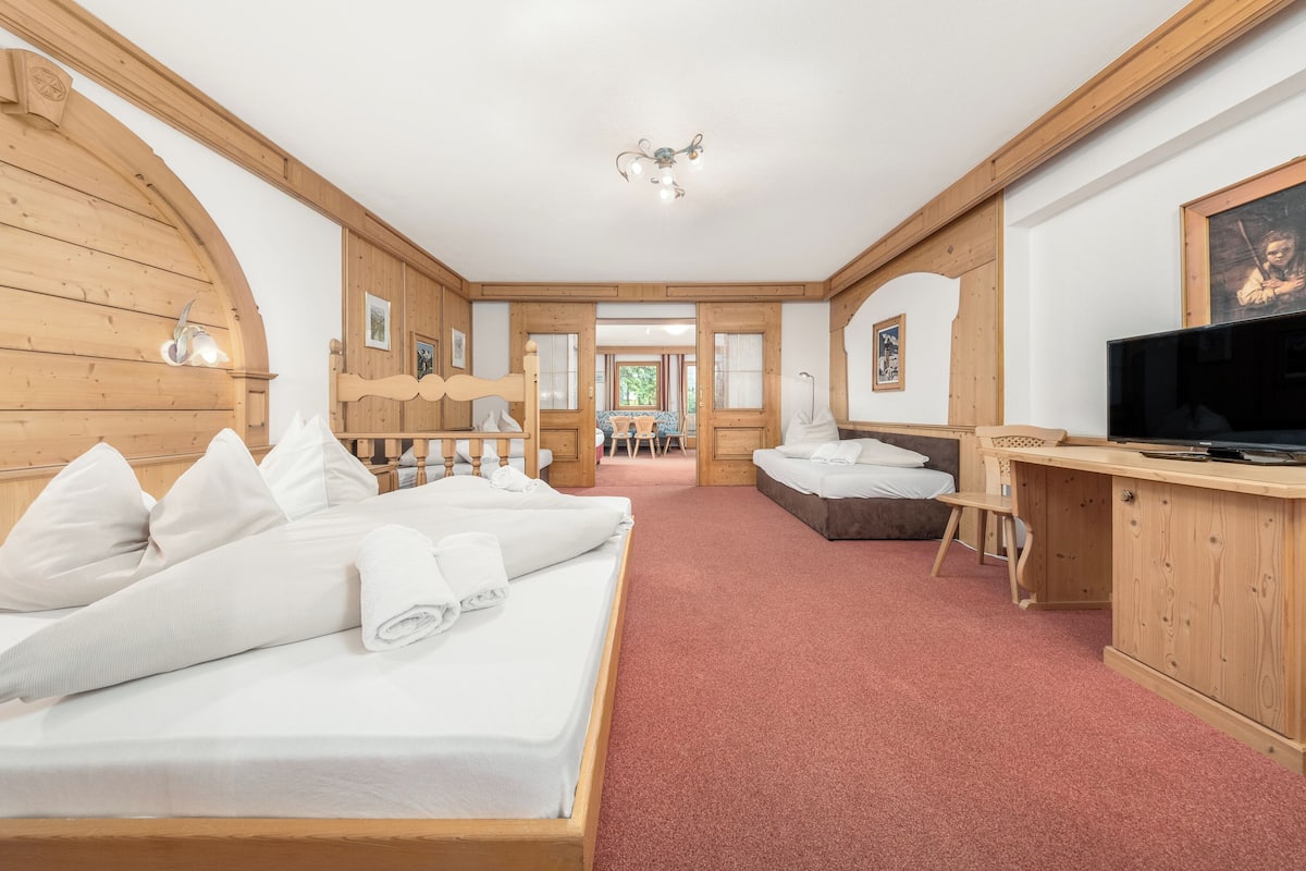 A hotel room with a wooden interior, a bed with white linens, a desk with a television, and a red carpet.