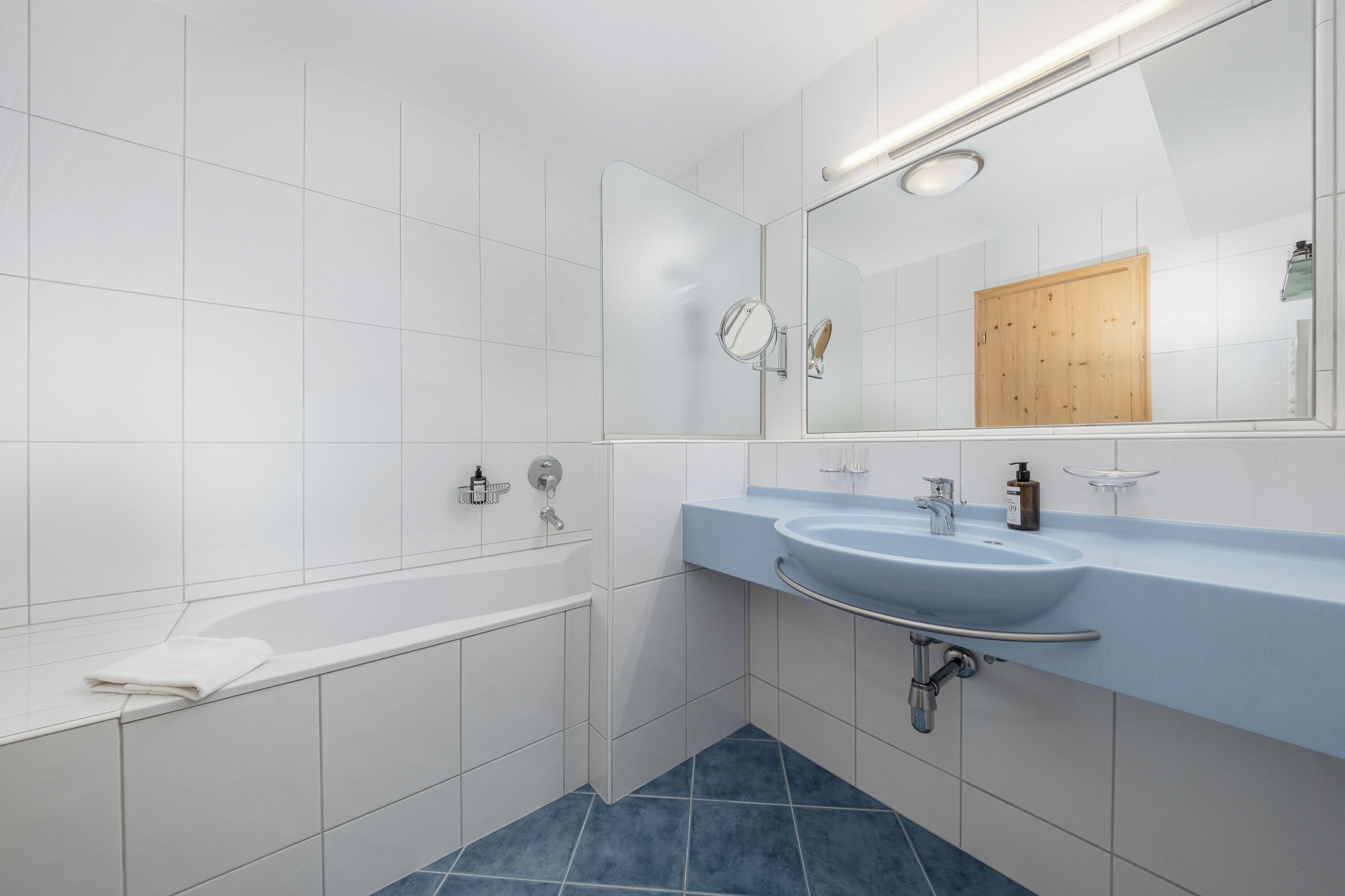 Ein modernes Badezimmer mit weiß gefliesten Wänden, blauem Fliesenboden, einer weißen Badewanne, einem Waschbecken mit Spiegel und einer Holztür.