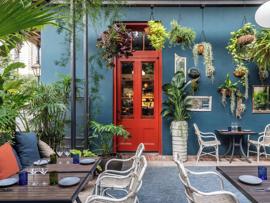 Uma área de refeições ao ar livre com cadeiras e mesas de vime, uma porta vermelha e diversas plantas penduradas.
