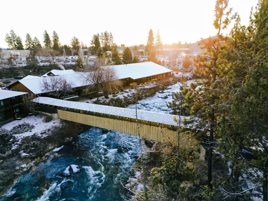 A wooden bridge over a river with snow-covered buildings and trees in the background.