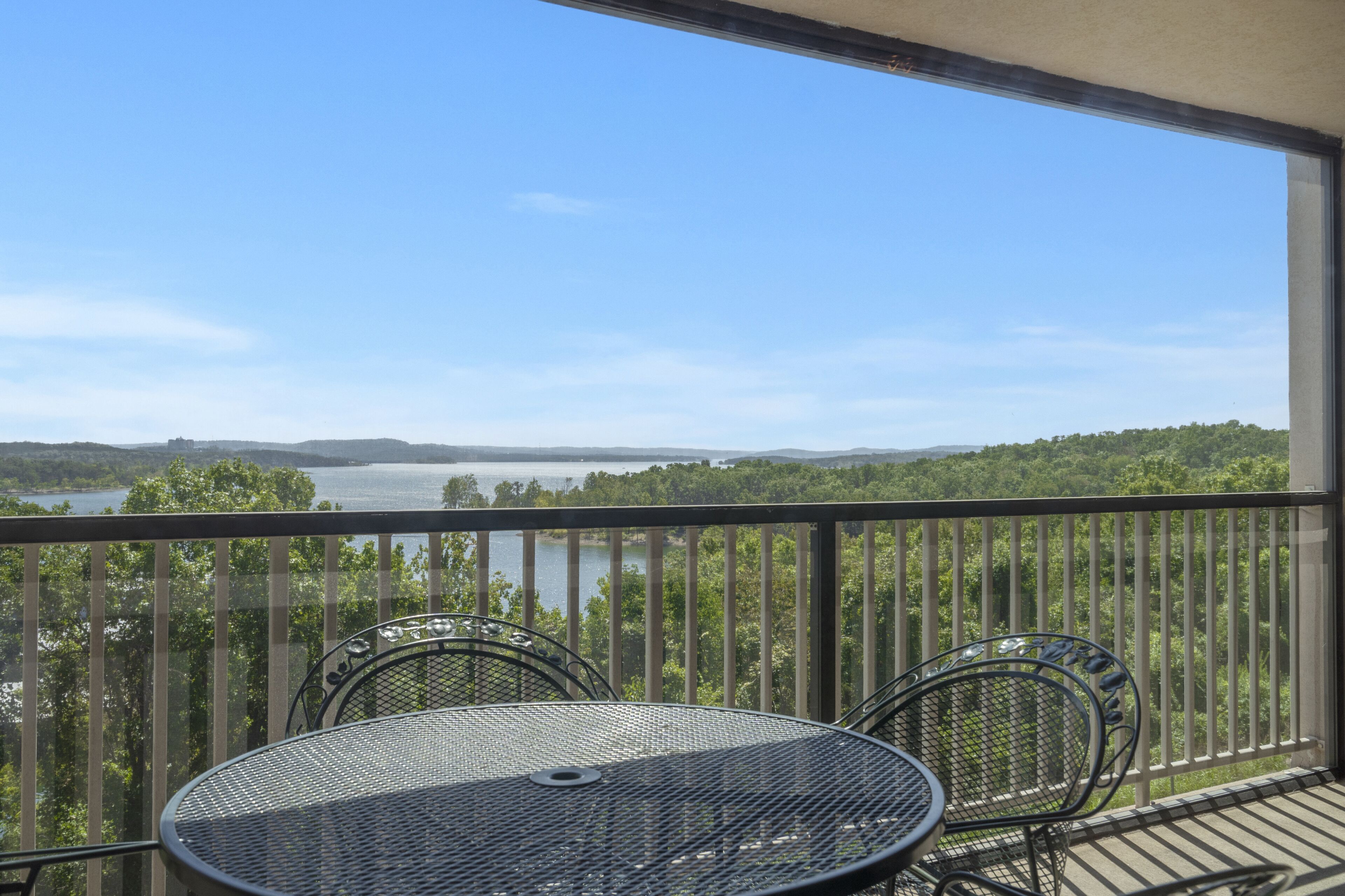 A balcony with a table and chairs, overlooking a lake and forested hills.