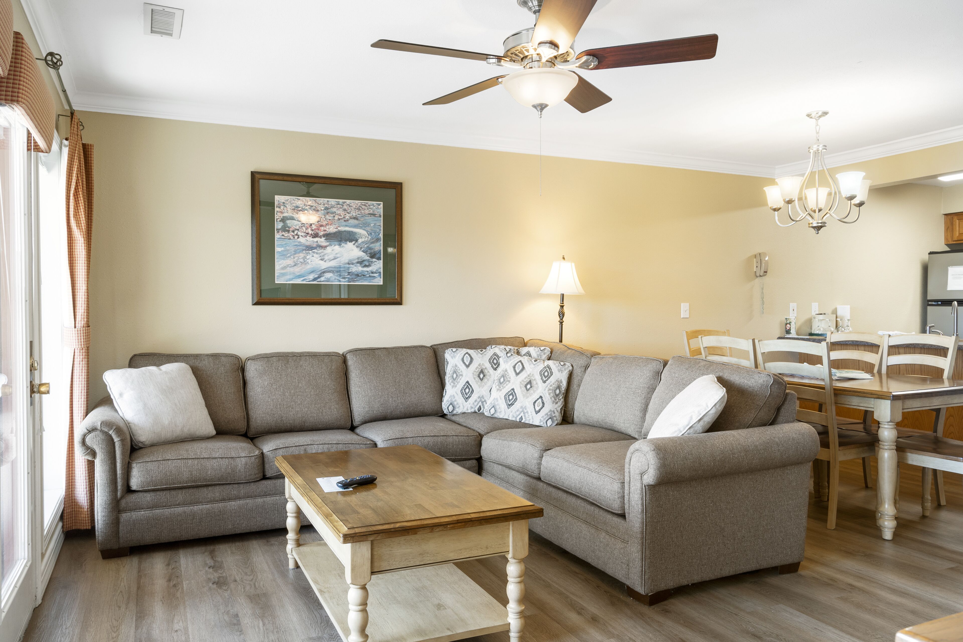 A living room with a sectional sofa, a wooden coffee table, a ceiling fan, a chandelier, and a dining area with a table and chairs.