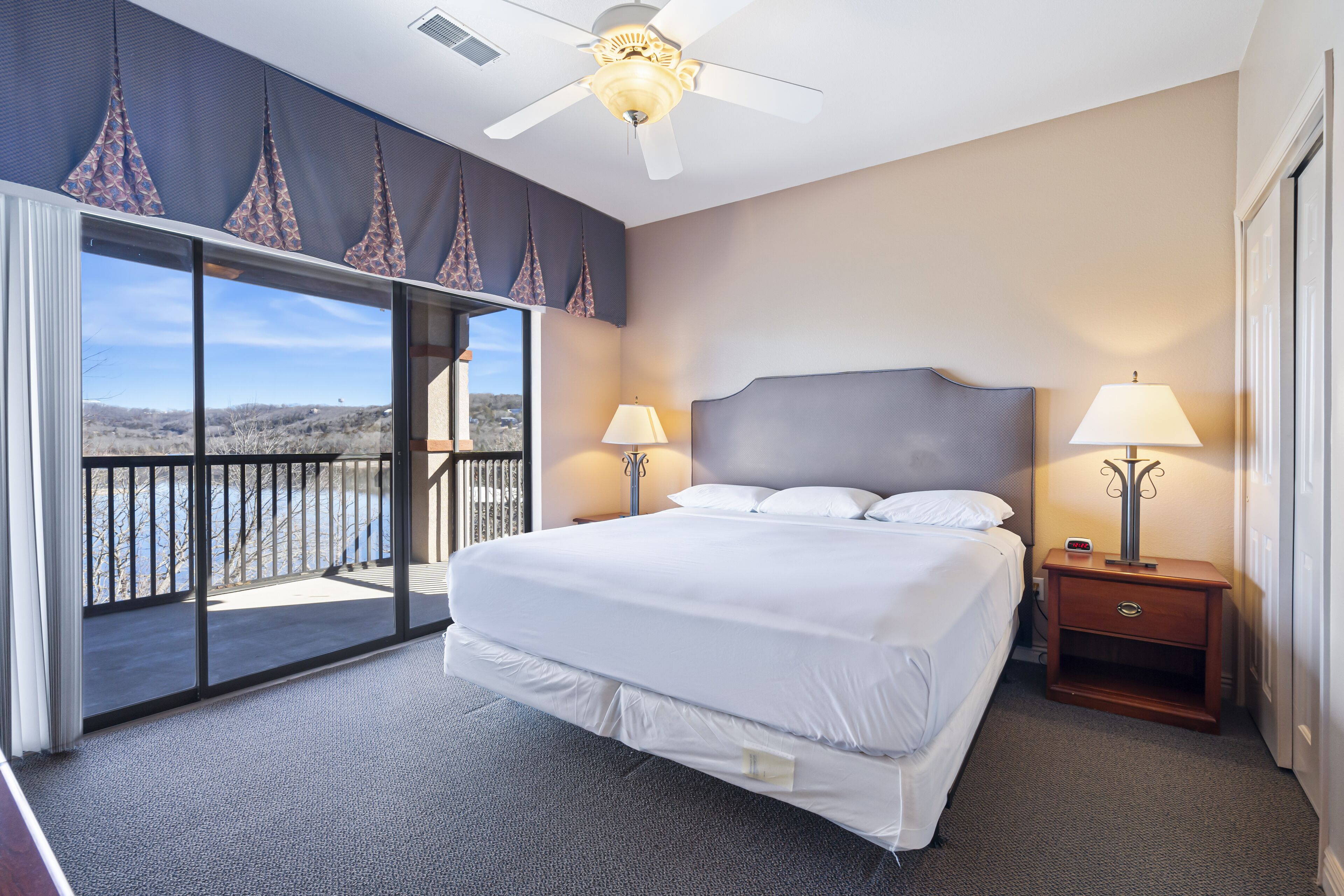 A hotel room with a large bed, two bedside lamps, a ceiling fan, and a view of a mountainous landscape through a sliding glass door.