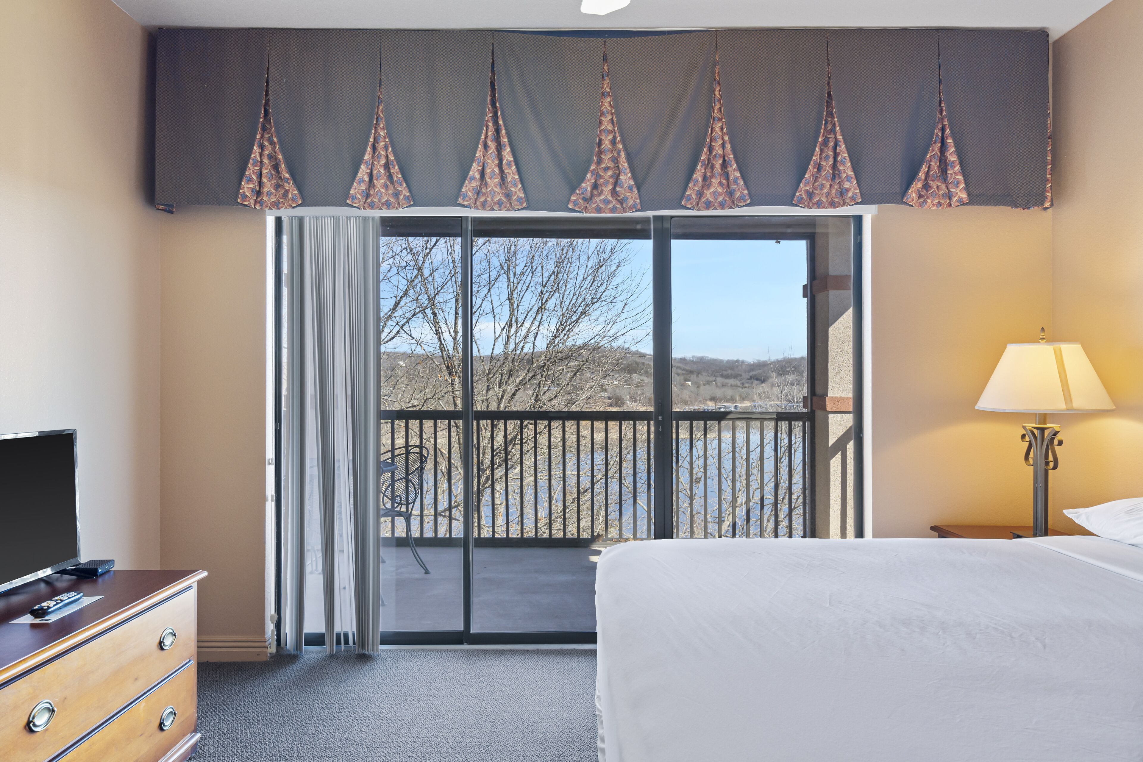 Une chambre d’hôtel comprenant un lit, une commode, une télévision, une lampe et un balcon avec vue sur le paysage.