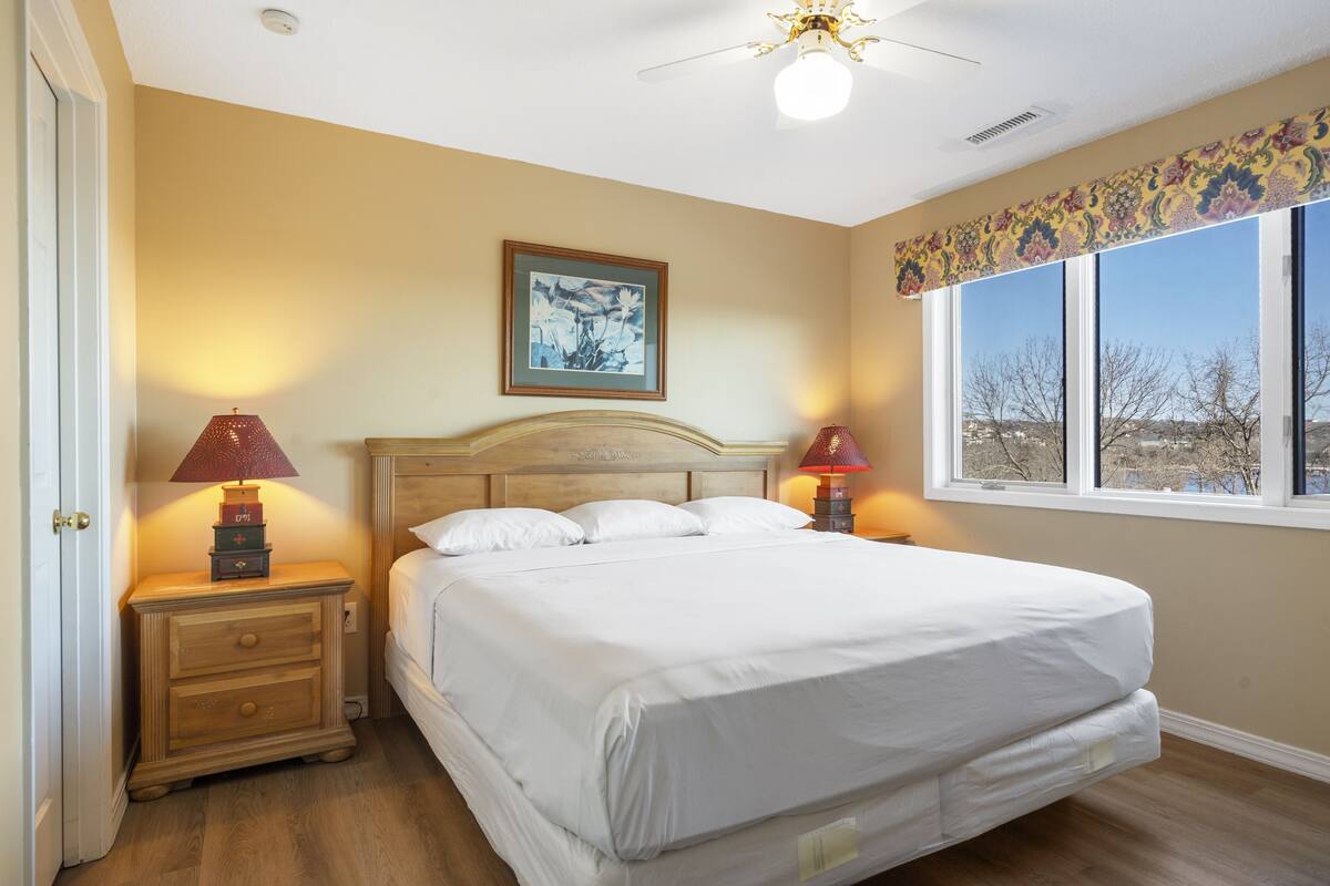 Une chambre avec un grand lit, deux tables de chevet, un ventilateur de plafond, une fenêtre avec vue et un tableau encadré au mur.