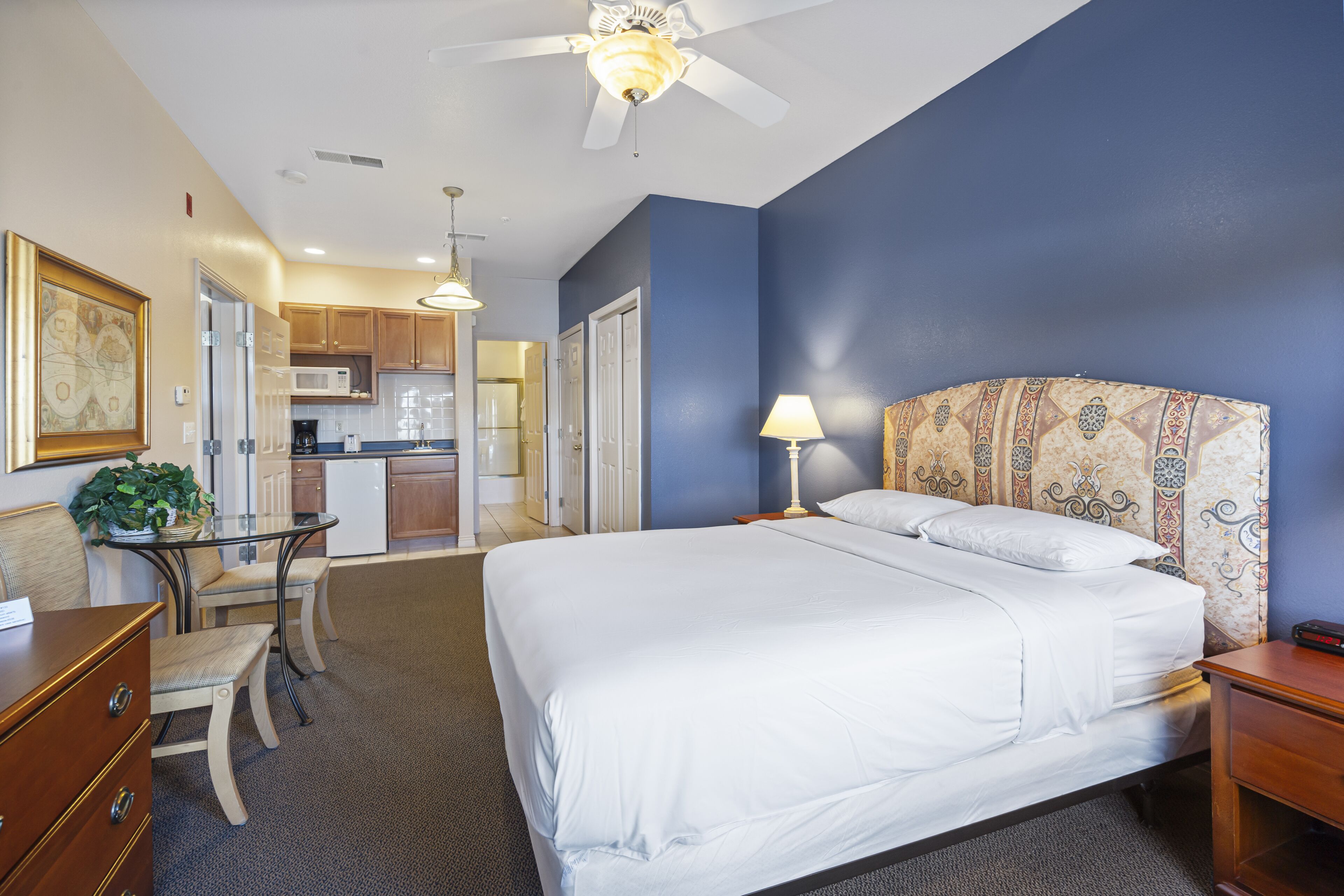 A hotel room with a large bed, a desk with a chair, a kitchenette, and a ceiling fan.