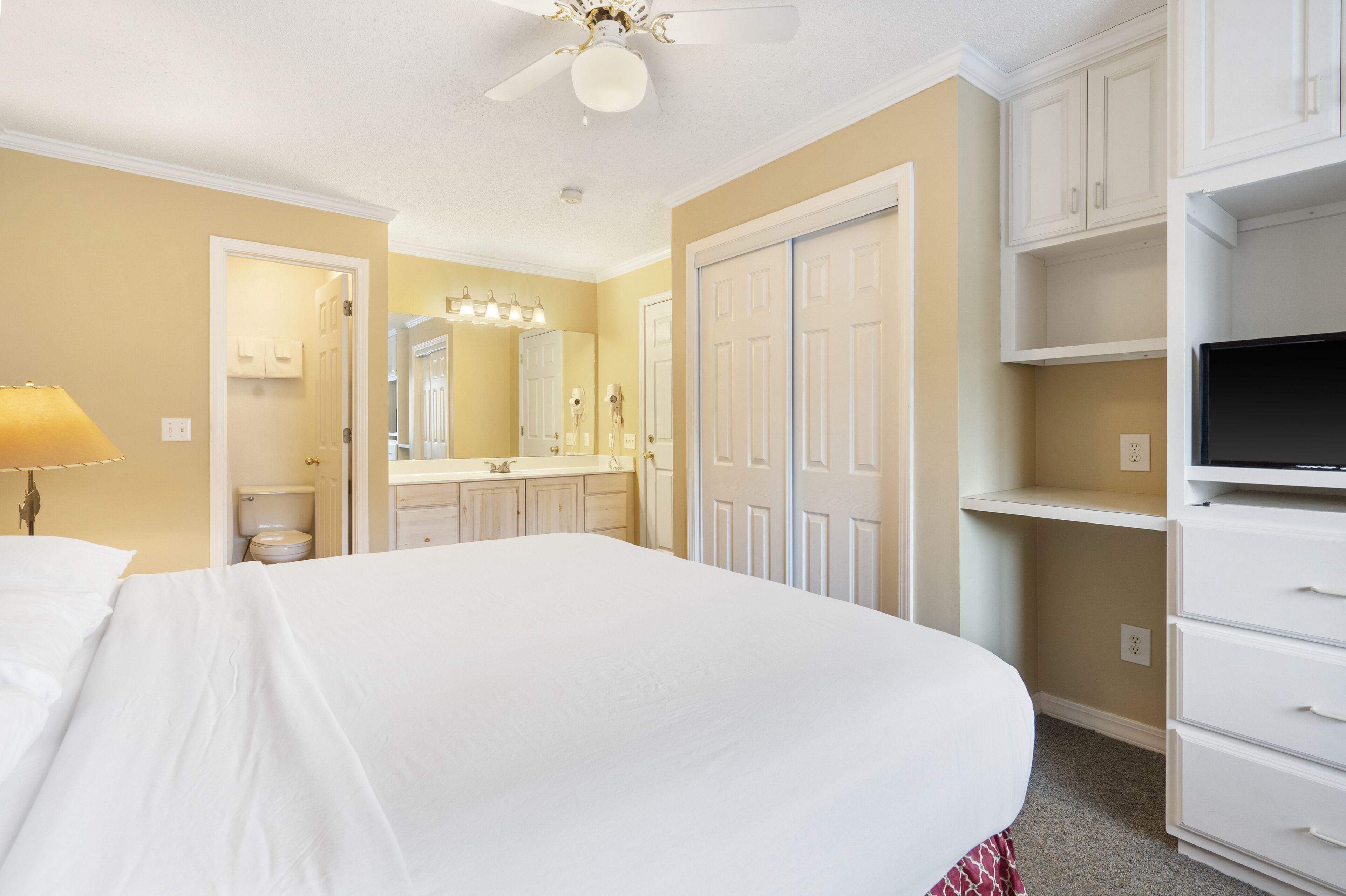 Une chambre d’hôtel avec un grand lit, une coiffeuse avec un miroir, un ventilateur de plafond et un téléviseur posé sur un meuble.