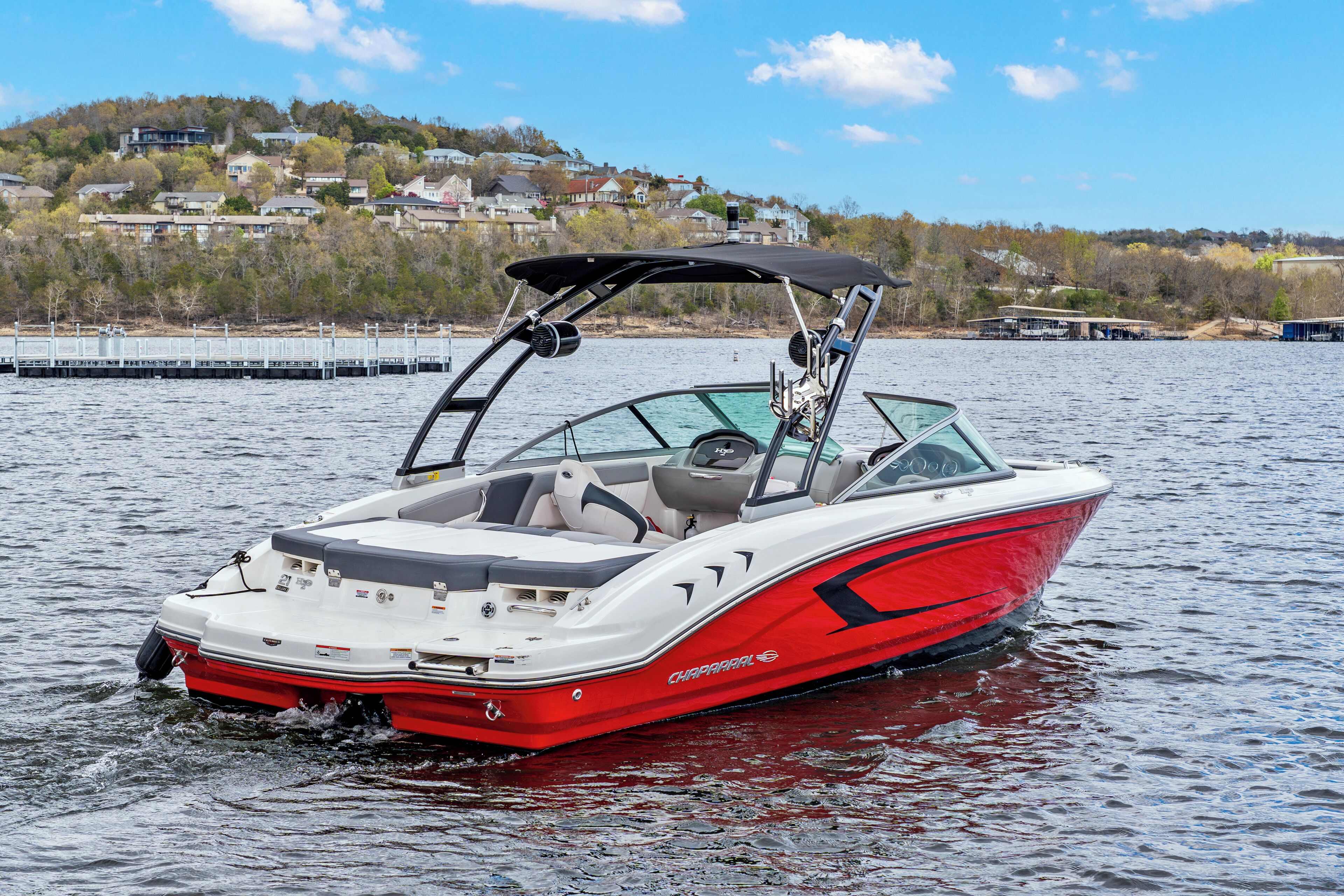 Un bateau à moteur rouge et blanc, doté d’un toit et de sièges pour les passagers, navigue sur un lac paisible, avec des maisons en arrière-plan.