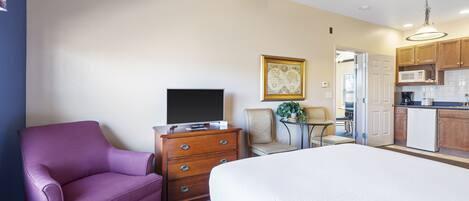Una habitación de hotel con cama, cómoda, televisor, silla, una mesita con una planta y una kitchenette al fondo.