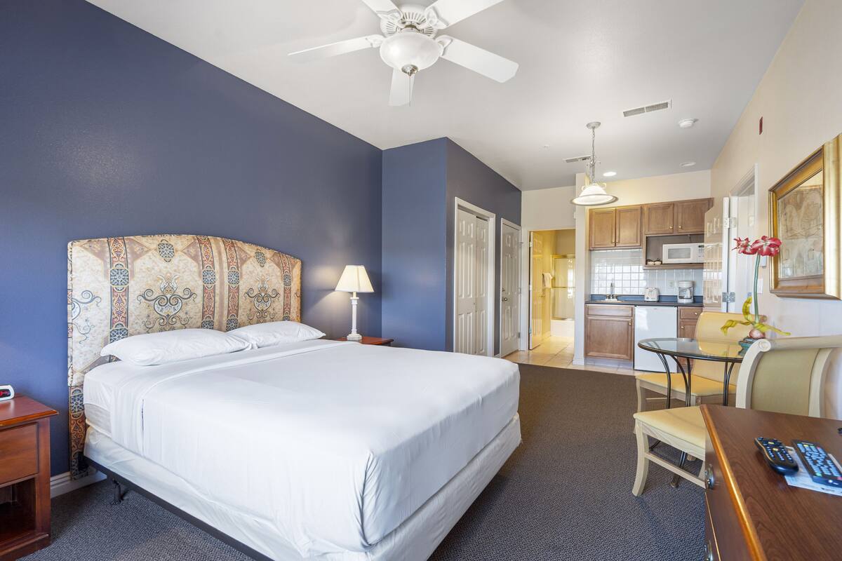 Une chambre d’hôtel avec un grand lit, un bureau avec une chaise, un ventilateur de plafond et, au fond, une petite cuisine.