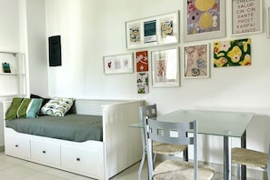 A room with a white sofa, a glass table, and chairs, a white radiator, and framed artwork on the wall. - M375 - Marcelli, two-room apartment with garden in a residence with swimming poo (Numana)