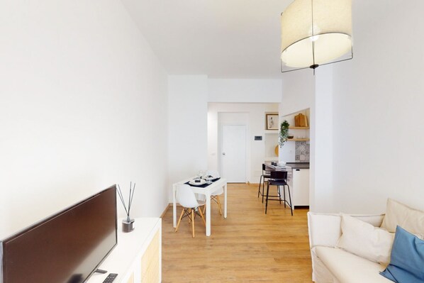A modern living room with a sofa, a dining table with chairs, a TV, and a kitchen area in the background.