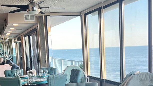 Un comedor con mesas dispuestas para cuatro, ventanas amplias que dan al océano y un ventilador de techo.