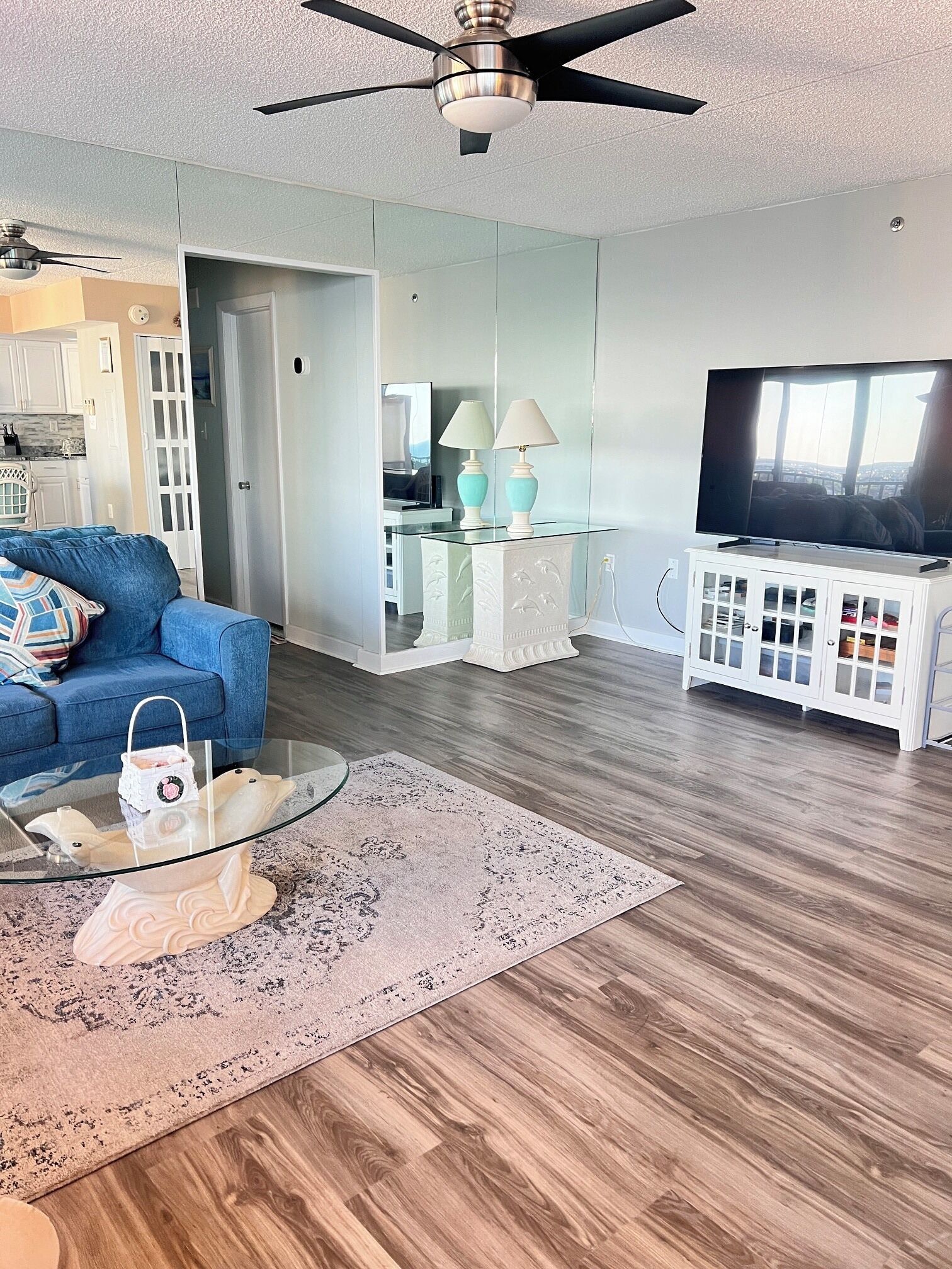 A modern living room with a glass coffee table, a blue sofa, a white cabinet with glass doors, a ceiling fan, and a television.