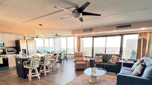 Una sala de estar moderna con comedor, ventilador de techo, ventanas amplias y vistas a la ciudad.