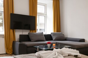 A modern living room with a sofa, a glass coffee table, a flat-screen TV mounted on the wall, and a window with curtains. - Spacious & Pleasant 2BR Apt. near Milenium Tower (Vienna)