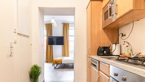 A narrow hallway leading to a bedroom with a bed, a potted plant, and a TV. The kitchen area includes wooden cabinets, a stove, and a kettle.