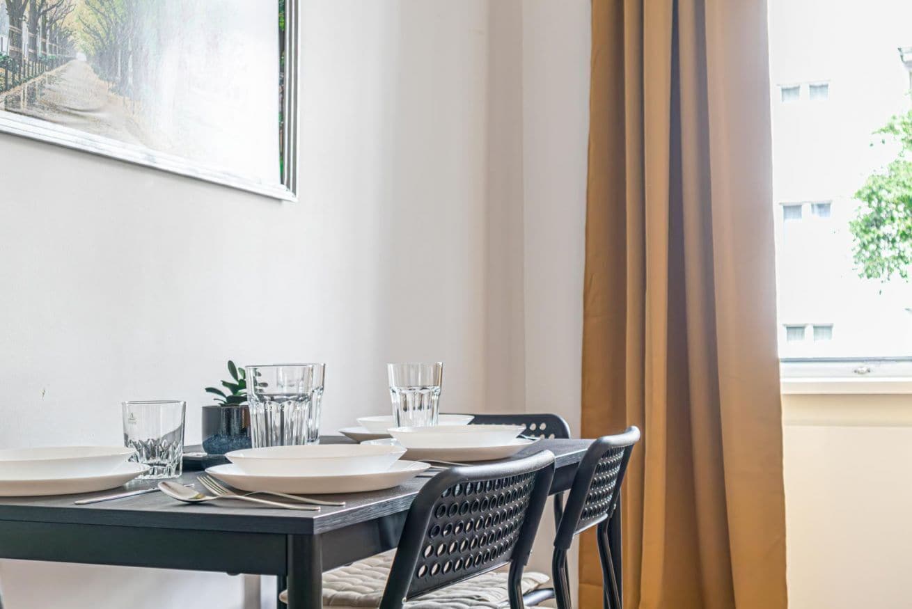 Una mesa de comedor con platos, vasos y cubiertos, una silla con respaldo de mimbre y una ventana con cortinas.