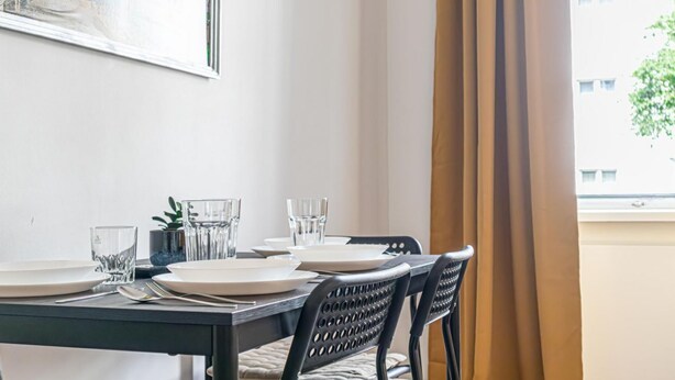 A dining table set with plates, glasses, and cutlery, a chair with a woven backrest, and a window with curtains.