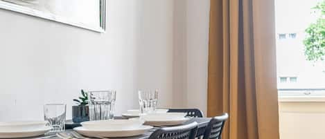 Una mesa de comedor con platos, vasos y cubiertos, una silla con respaldo de mimbre y una ventana con cortinas.